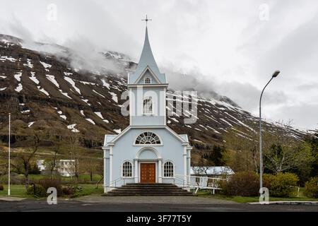 Symmetrische Frontansicht der blauen Holzkirche Seyðisfjarðarkirkja vor bewölkten Bergen in Seyðisfjörður in Ostisland Stockfoto