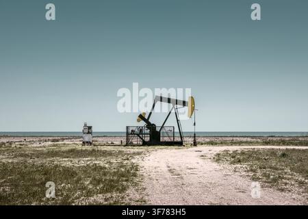 Öl-Pumpjack und Bohrkopf in einer spärlichen ländlichen Landschaft unter einem klaren blauen Himmel mit reichlich Kopierraum. Stockfoto