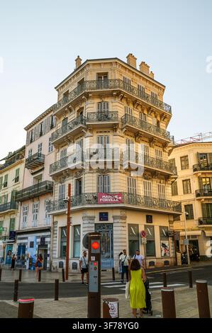 Cannes, Frankreich - 29. Juli 2026: Ein mehrstöckiges Gebäude mit verzierten Balkonen und geschlossenen Fensterläden steht an einer Straßenecke mit Fußgängern. Stockfoto