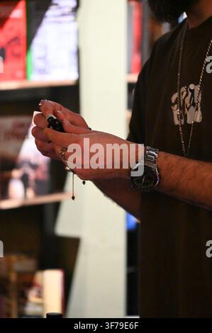 Eine Nahaufnahme, die die Hände eines DJs zeigt, die mit silbernen Ringen und einer Uhr geschmückt sind, während er sorgfältig die empfindliche Nadel auf einer Tonarm-Kartusche eines Plattenspielers einstellt. Der Plattenspieler spielt eine Platte ab, und im Hintergrund sind verschwommene Vinyl-Regale zu sehen. Stockfoto