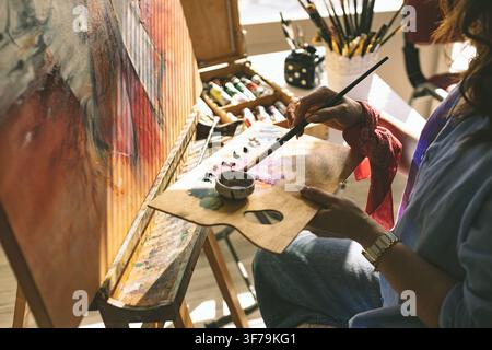 Nahaufnahme einer Künstlerin, die Farbe auf einer Palette mischt, während sie an einer Original-Leinwand in einem hell erleuchteten Atelier arbeitet. Stockfoto