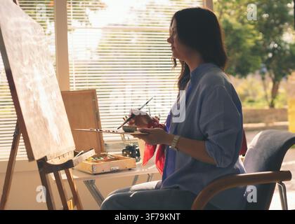 Seitenansicht einer Künstlerin, die auf einer Leinwand in einem hellen Atelier bei natürlichem Sonnenlicht malt, mit Kunstutensilien. Stockfoto