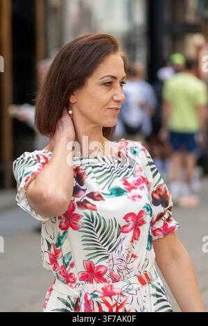 Eine Frau in einem farbenfrohen Blumenkleid steht im Freien, während sie mit der Hand ihren Hals berührt und zur Seite schaut. Stockfoto