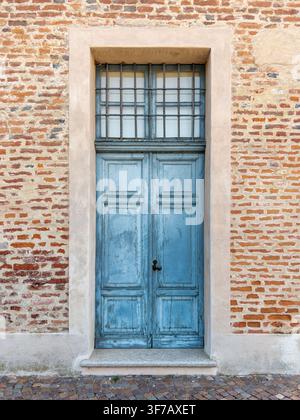 Alte verwitterte blaue Doppeltür aus Holz mit eisernen Sicherheitsgittern am Fenster, eingebettet in einer historischen roten Backsteinmauer in Italien. Vintage-architektonisches D Stockfoto