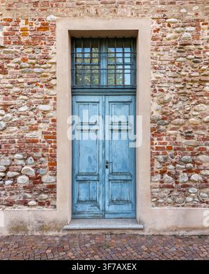Alte blau bemalte Holztür, eingebettet in einer traditionellen rustikalen Wand aus roten Ziegeln und Flusssteinen. Die verwitterte Tür zeigt dekorative Paneele Stockfoto