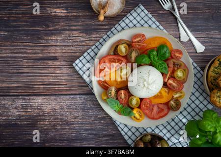 Frische bunte Tomaten serviert mit cremiger Burrata-Käse und Basilikum auf einem rustikalen dunklen Holztisch mit geschnittenem Brot und mediterranem Stil Stockfoto