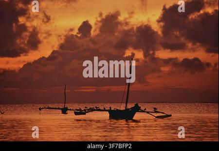 Zwei traditionelle Holzboote liegen im ruhigen Wasser vor Diani Beach während eines intensiven Sonnenaufgangs. Der Himmel leuchtet in warmen Orangetönen und Rot, dunkle Wolken Stockfoto