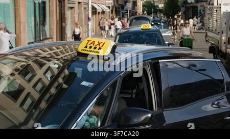 STRASBOURG, FRANKREICH - 6. AUGUST 2026 - Taxi mit Straßburg-Schild fährt eine belebte Stadtstraße mit Menschen und Verkehr Stockfoto