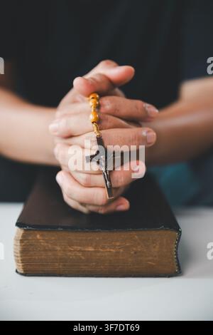 Nahaufnahme von verschlungenen Händen, die Rosenkranzperlen und ein Kruzifix über einer alten Heiligen Bibel halten, was katholisches Gebet, christlichen Glauben, Hingabe, Hoffnung darstellt, Stockfoto