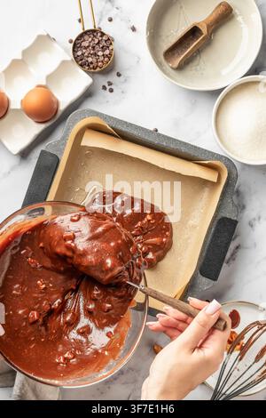 Prozess der Herstellung von Schokoladenbrownie. Weibliche Hände gießen den Teig in eine Backform, Ansicht von oben. Konzept für Heimbäckerei und Kulinarik, Rezept Stockfoto