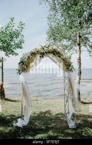 Eleganter floraler Hochzeitsbogen, geschmückt mit weißen Blumen, Grünpflanzen und fließendem transparentem Stoff, aufgestellt an einem grasbewachsenen Ufer am Meer unter Birken, kreati Stockfoto