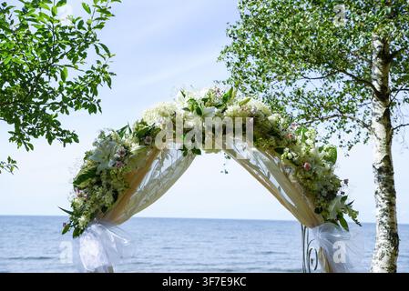 Eleganter Hochzeitsbogen geschmückt mit weißen Blumen, üppigem Grün und fließendem transparentem Stoff, eingerahmt von Bäumen vor einem ruhigen Meereshintergrund, der eine Romantik schafft Stockfoto