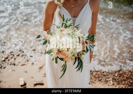 Elegante Braut, die einen romantischen pastellfarbenen Hochzeitsstrauß an einem sonnenbeschienenen Strand hält, der Küstenlandschaft, Brautmode und die Ästhetik von Destinationsehen kombiniert Stockfoto