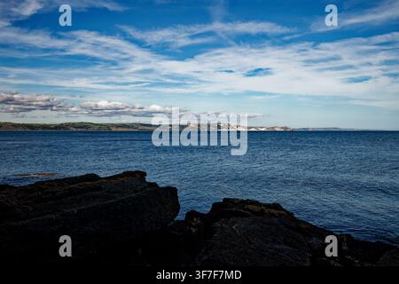 Ein ruhiges blaues Meer plätschert sanft gegen Uferfelsen mit einer Küstenlinie aus weißen Kreidefelsen in der Ferne, Weymouth Bay, Dorset, England. Stockfoto
