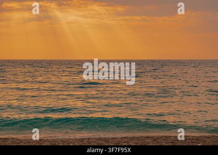 Sonnenstrahlen, die durch die Wolken über ruhigen Meereswellen an der sandigen Strandküste während eines dramatischen orangefarbenen Sonnenuntergangs in Lefkada, Griechenland, für eine Naturlandschaft Stockfoto