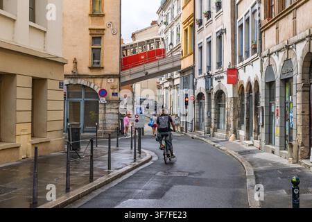Lyon, Frankreich – 27. Juli 2026: Straßenansicht im Vieux Lyon mit einem Radfahrer, Fußgängern, historischen Fassaden und der roten Standseilbahn-Brücke darüber. Stockfoto