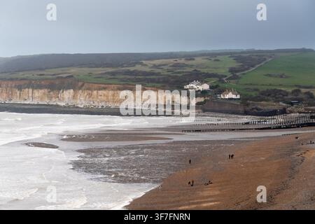 Besucher spazieren am Strand von Cuckmere Haven und genießen die Aussicht auf die historischen Coastguard-Cottages und die ikonischen Seven-Sisters-Klippen in East Sussex. Stockfoto
