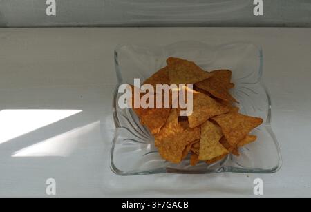 Nacho-Tortilla-Chips füllen eine Glasschale und bieten einen knusprigen und salzigen Snack auf einer weißen Oberfläche, wobei natürliches Fensterlicht einen Schatten wirft Stockfoto