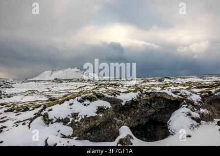 Eine kleine Lavahöhle öffnet sich zwischen vulkanischen Felsen, die im Vordergrund mit Schnee und Moos bedeckt sind, mit einem weiten Lavafeld und einem schneebedeckten Vulkan im Hintergrund Stockfoto