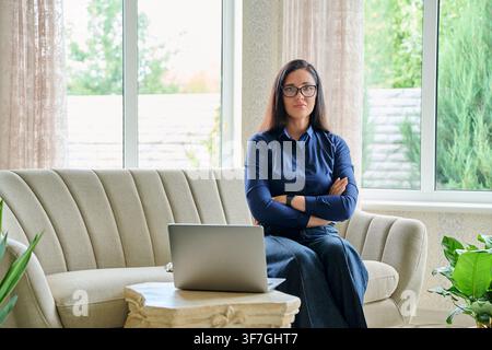 Selbstbewusste Geschäftsfrau mit Laptop, die auf dem Sofa im heimischen Remote-Arbeitsplatz sitzt Stockfoto