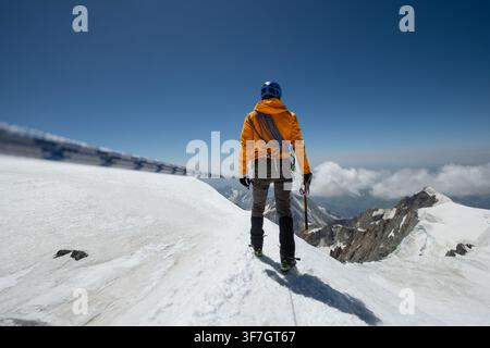 Bergsteiger in orangefarbener Jacke hält Eispickel, steht auf schneebedecktem Grat, fokussierter Schuss Mont Blanc nahe der Vallot-Hütte, Hochgebirgs-Expedition in den französischen Alpen Stockfoto