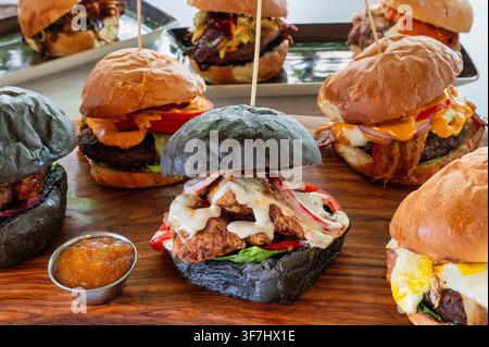 Sortiment von Gourmet-Burgern, die auf einem Holzbrett serviert werden, mit schwarzen und traditionellen Brötchen, Rind- und Hühner-Patties, geschmolzenem Käse, Gemüse, Bac Stockfoto
