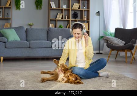 Junge Frau mit Kopfhörern streichelt einen Cocker Spaniel auf dem Teppich zu Hause im Wohnzimmer Stockfoto