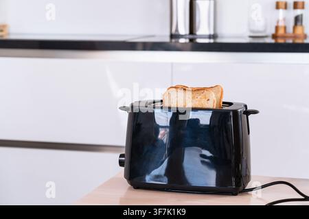 Zwei Brotscheiben steigen auf, rösten und bräunen sich in einem schwarzen elektrischen Toaster auf einer modernen Küchentheke, Zubereitung des Frühstücks, Hausmannskost, Heimfrühstück Stockfoto