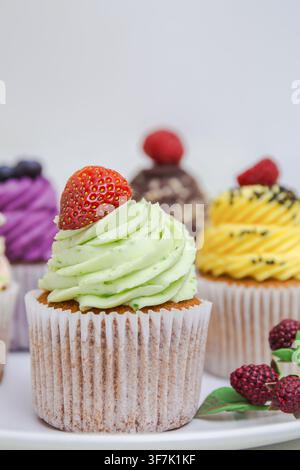 Farbenfroher Cupcake mit hellgrün geschwungener Glasur und frischer Erdbeere, umgeben von verschiedenen dekorierten Cupcakes und Beeren, natürliche Färbung Stockfoto