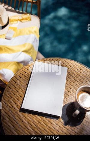 Das leere Notizbuch liegt auf einem Korbtisch neben einer Tasse Kaffee und einem gelb gestreiften Handtuch, und im Hintergrund ist ein Swimmingpool zu sehen Stockfoto