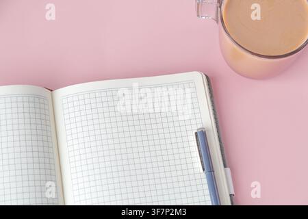 Ein offenes Gitter-Notizbuch mit einem Stift und einer Tasse Kaffee auf einem rosa Hintergrund. Das minimalistische Arbeitsplatz-Setup repräsentiert Fokus und Produktivität. Konzept von Studium und Planung Stockfoto