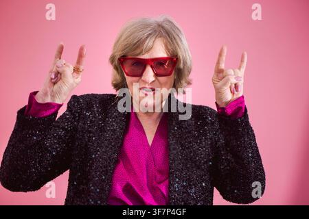 Eine ältere weiße Frau mit kurzen Haaren und roter Sonnenbrille posiert mit einer Geste und trägt eine schwarze Paillettenjacke vor einem leuchtend rosa Hintergrund Stockfoto