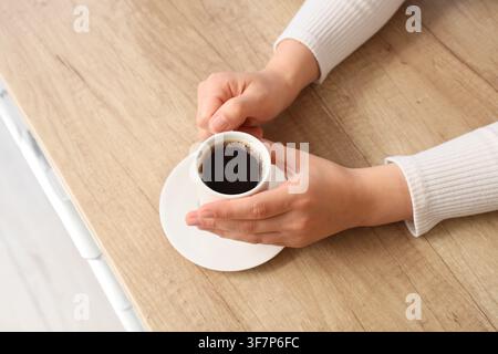 Frauenhände halten eine kleine Tasse schwarzen Kaffees auf einer weißen Untertasse über einem hellen Holztisch, Blick von oben Stockfoto