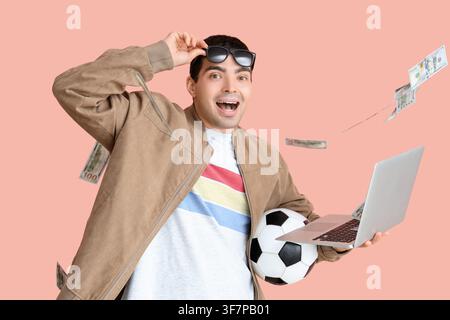 Überraschter junger Mann mit Fußball, Laptop und fallendem Geld auf rosa Hintergrund. Sportwettenkonzept Stockfoto
