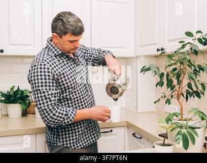 Ein Mann gießt heißes Wasser aus einem Wasserkocher in eine weiße Tasse, um Tee oder Kaffee zuzubereiten. Positives Lebensgefühl Stockfoto