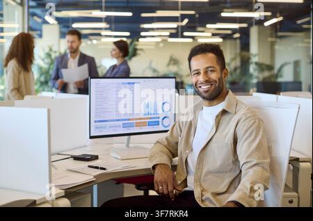 Lächelnder Finanzanalyst am Schreibtisch mit Daten-Dashboard und Diagrammen im Büro Stockfoto