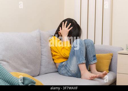 Krankes asiatisches Mädchen fühlt sich unwohl, sitzt auf dem Sofa, Kindheitserkrankung, Schmerzkonzept Stockfoto