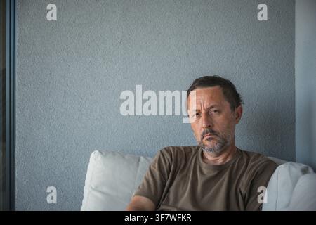 Ernster Mann mittleren Alters sitzt auf einem weißen Sofa vor einer neutralen grauen Wand, wirkt nachdenklich und müde. Legere Kleidung und sanftes natürliches Licht vermitteln s Stockfoto