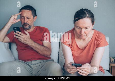 Mittelalterliches Paar, das Smartphones benutzt, während es zusammen im Freien sitzt, wirkt gelangweilt und in getrennte Bildschirme vertieft. Beziehungsstress, Technologie Stockfoto