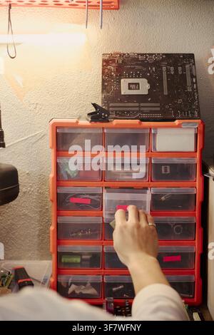 Hand einer jungen asiatischen Frau, die in einer Tech-Workshop eine kleine Plastikschublade in einem Organizer öffnet und elektronische Komponenten für den Hardware-Reparaturdienst sortiert, vertikal Stockfoto