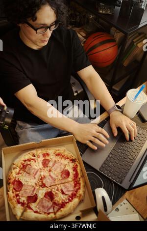 Vertikales Bild eines jungen asiatischen Mannes, der auf einer Laptop-Tastatur tippt, neben einer offenen Pizzaschachtel, während er von zu Hause aus arbeitet und zu Mittag isst, geeignet für Marketing von Telearbeit und Lebensmittellieferung Stockfoto