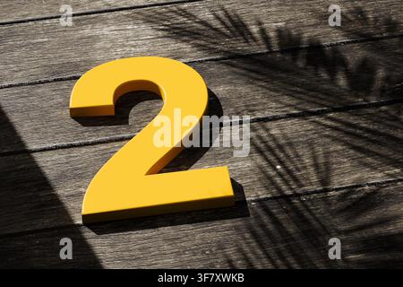 Gelbe Zahl zwei auf dunklen Holzplanken, mit dem Schatten einer tropischen Palme, der ein Outdoor-Muster erzeugt Stockfoto