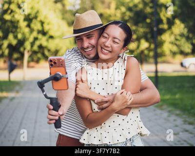 Junger Mann umarmt seine Freundin von hinten, während sie einen verspielten Moment zusammen auf einem Smartphone einfangen. Stockfoto