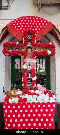 Ein mit Blumen bedecktes Mai-Kreuz mit einer Darstellung farbenfroher dekorativer Schädel steht vor einem grünen Fenster, eingerahmt von roten und weißen Blumen Stockfoto