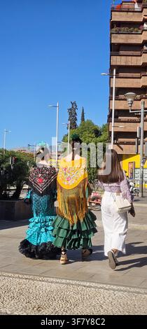 Drei Frauen gehen durch Granada, zwei tragen farbenfrohe traditionelle andalusische Kleider mit gerüschten Röcken, floralen Haaraccessoires und bestickten Fri Stockfoto