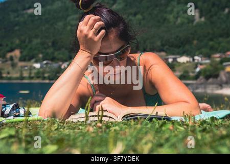 Eine Frau liest ein Buch, während sie auf einer Wiese nahe dem Ufer des Lago di Piazze im Trentino liegt. Die Frau hat langes dunkles Haar und eine Sonnenbrille im Gesicht Stockfoto
