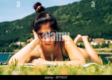 Eine Frau liest ein Buch, während sie auf einer Wiese nahe dem Ufer des Lago di Piazze im Trentino liegt. Die Frau hat langes dunkles Haar und eine Sonnenbrille im Gesicht Stockfoto