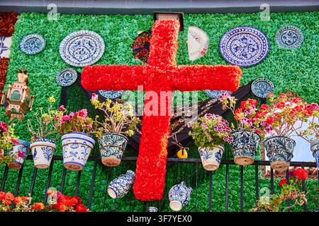 Eine große rote Blume bedeckte Kreuz beherrscht eine aufwendige Cruces de Mayo-Ausstellung mit bemalten Keramiktopfen, dekorativen Tellern, Geranien, einer Stickerei Stockfoto