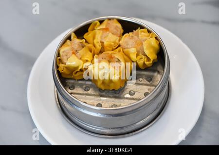 Hochwinkelschuss von vier gedämpften gelben Schweine- und Garnelen-Shumai-Dumplings, serviert in einem traditionellen Metall-Dampfgarer auf einem weißen Teller. Chinesisches Dim Sum Stockfoto