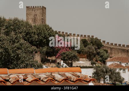 Ein steinerner Wachturm und zinnenbewehrte Mauern der Burg Óbidos erheben sich über Bougainvillea und Dächern in Óbidos, Portugal, am 27. Juli 2026. Stockfoto